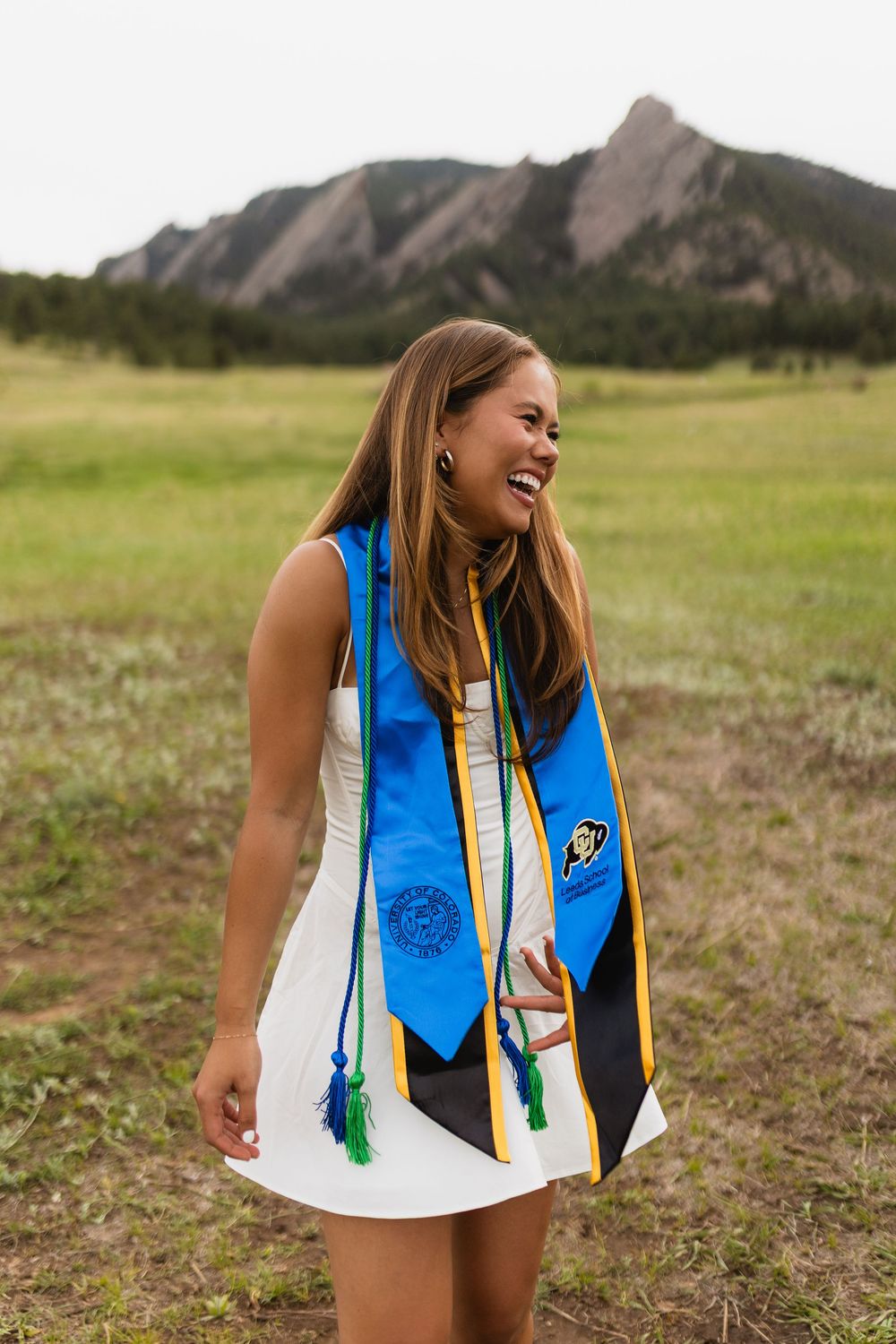 Graduate in white dress and blue graduation stoles smiles joyfully with the Flatirons mountains of Boulder in background.