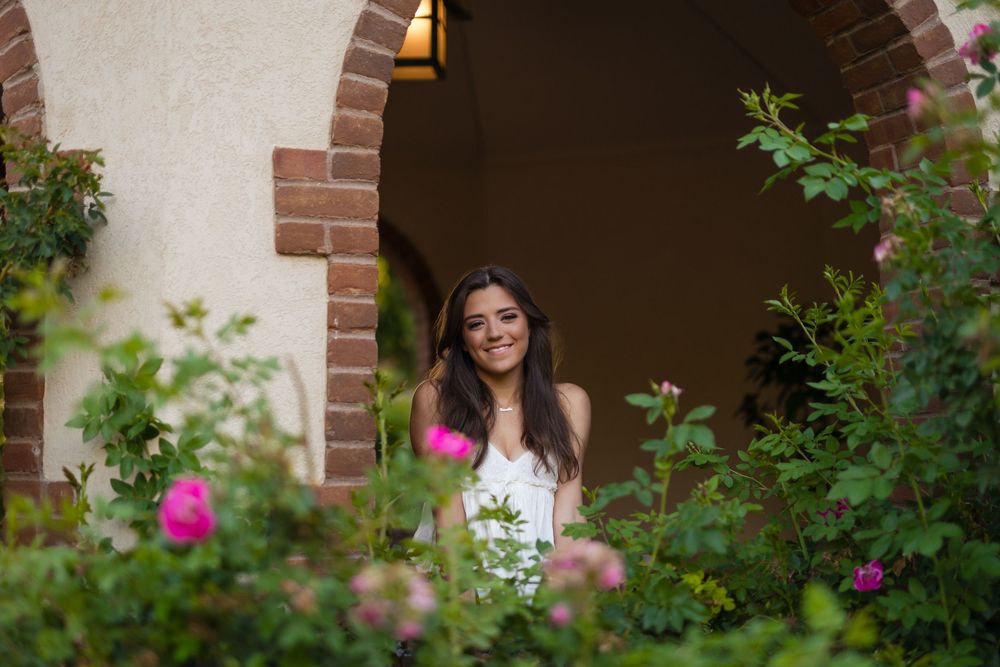 Smiling young woman standing in a brick archway surrounded by lush green foliage and pink roses in a garden.