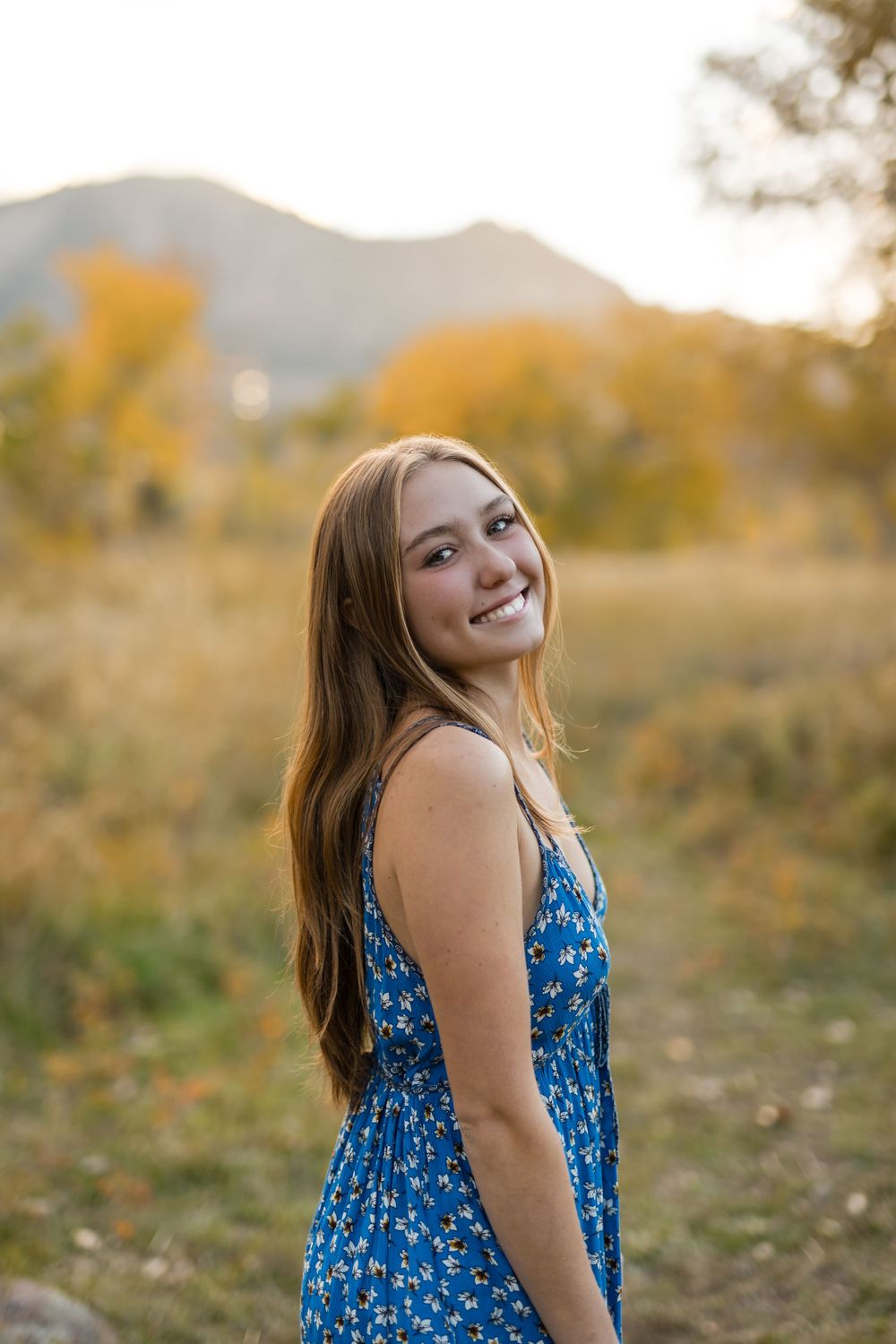 Person in a blue floral dress poses outdoors during golden hour with natural scenery in the background.