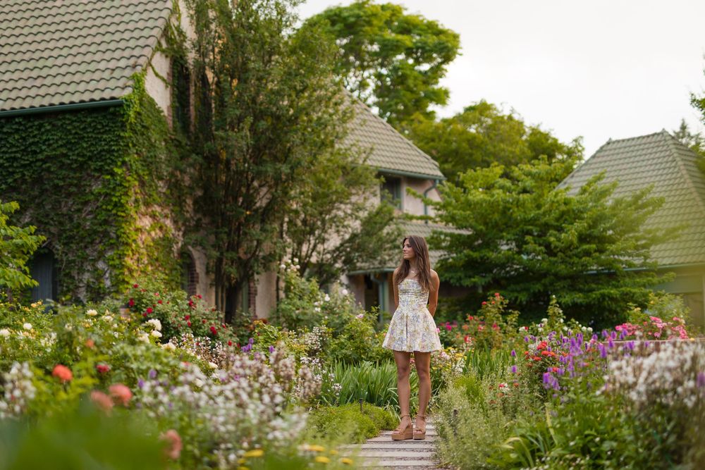 Woman in white dress standing in a lush cottage garden with colorful flowers and ivy-covered stone house.