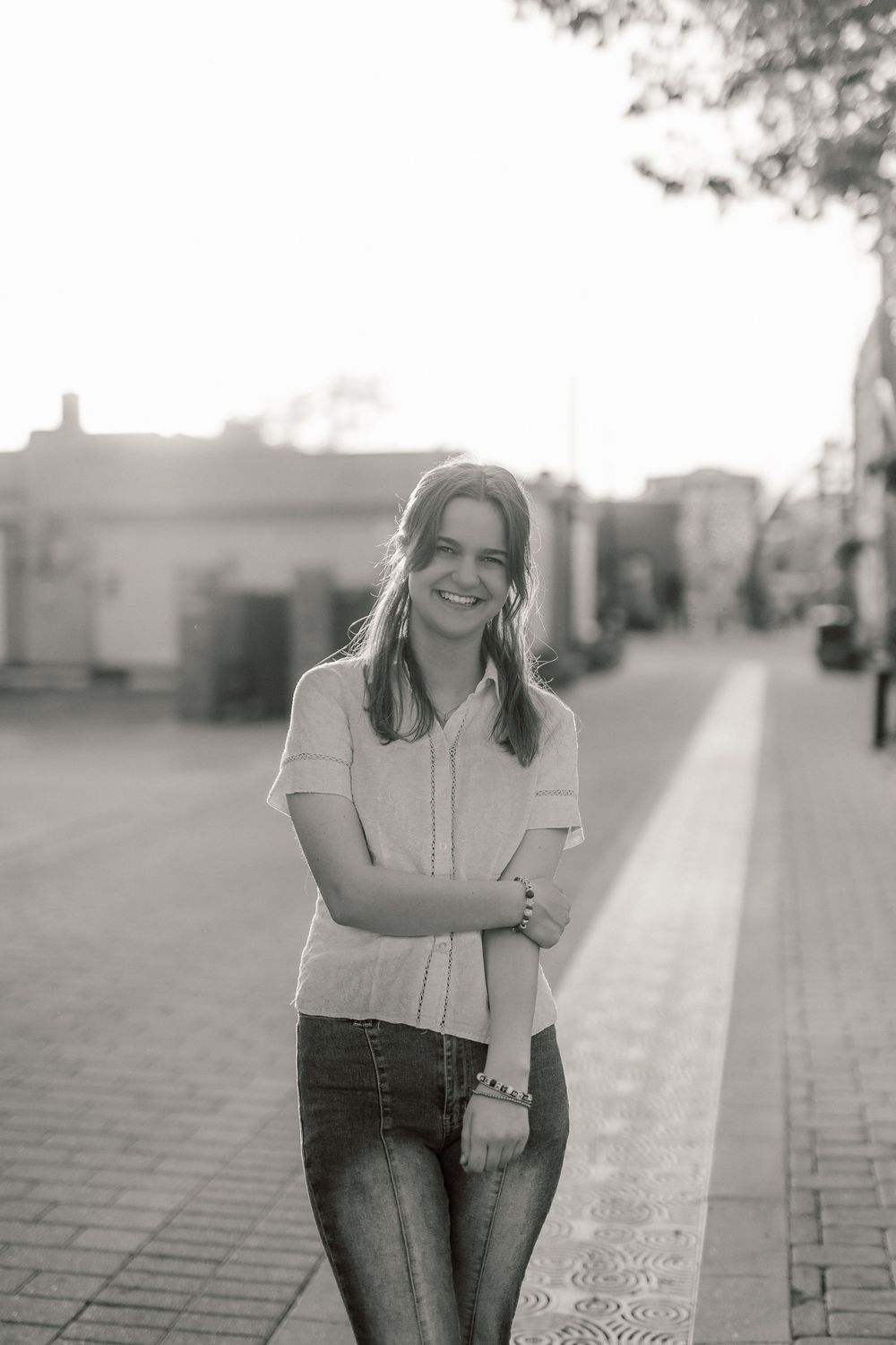Black and white photo of a smiling young woman in jeans and blouse standing on a sunlit urban sidewalk.