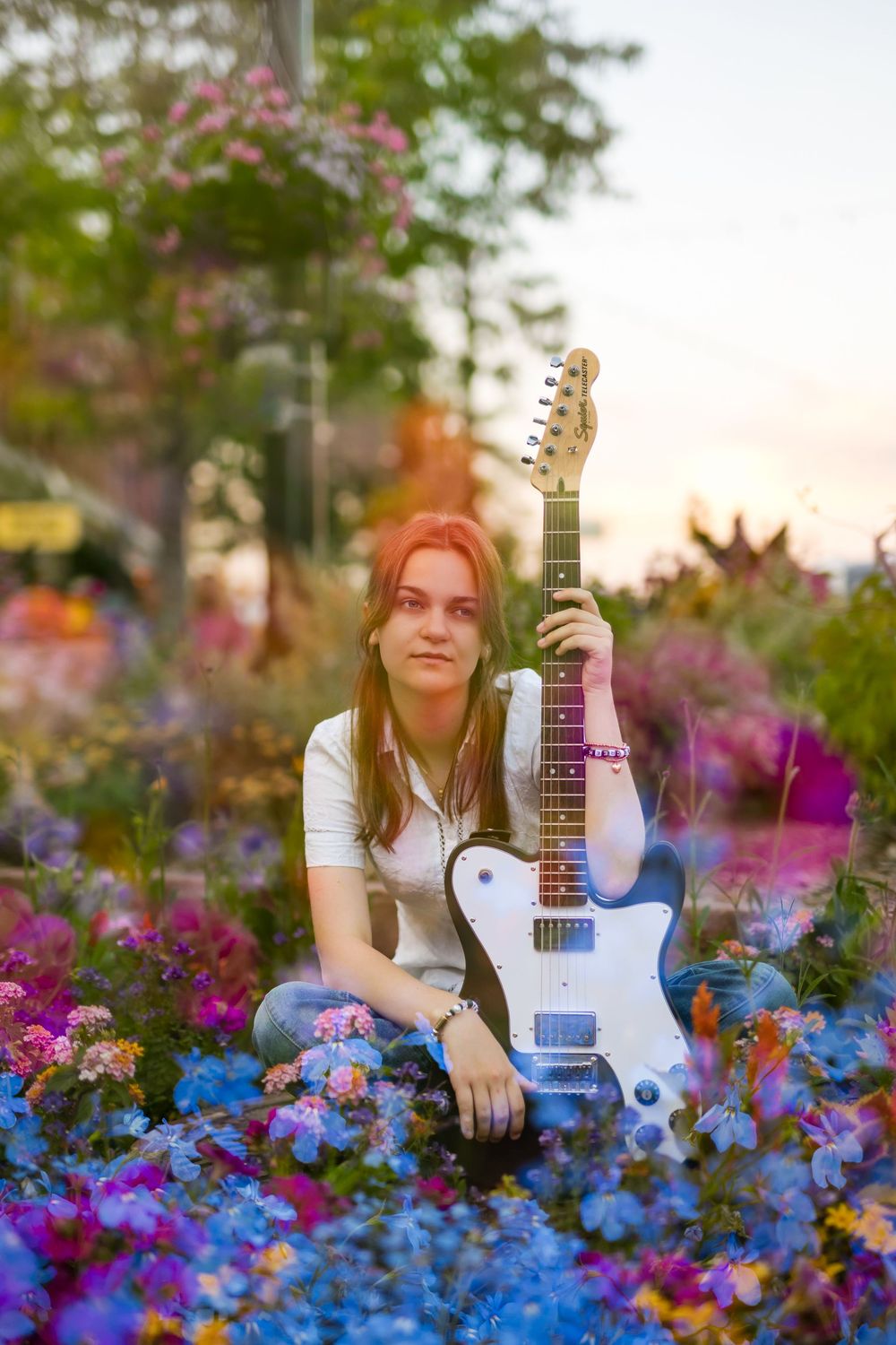 Young woman sitting among colorful flowers holding a white electric guitar at sunset.