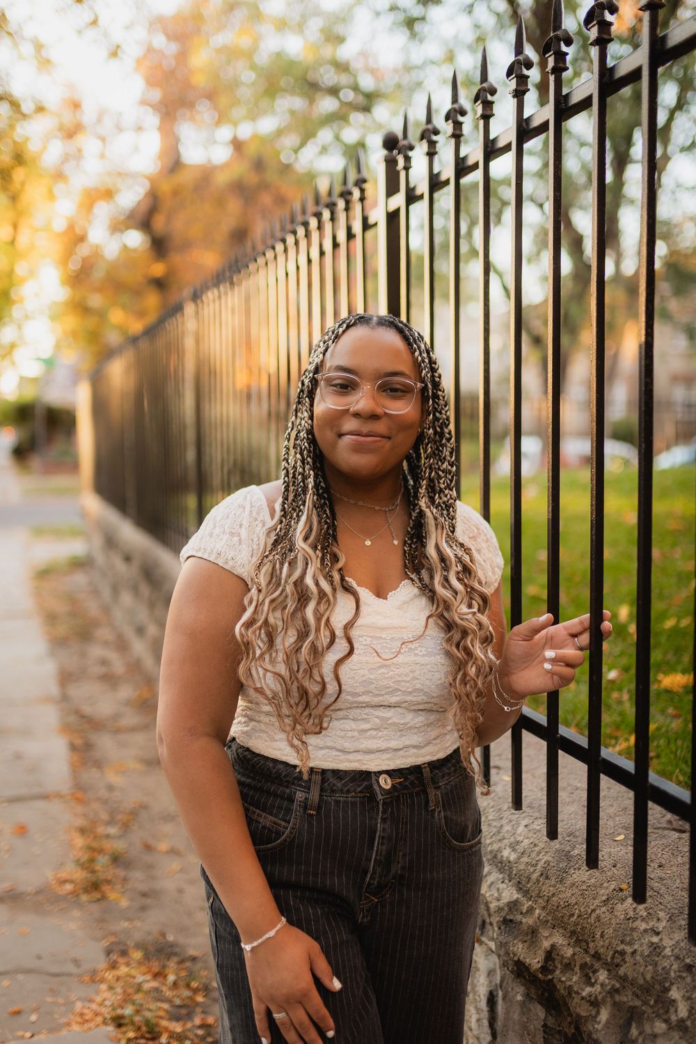 A person poses near an iron fence on an autumn day with fallen leaves and warm lighting.