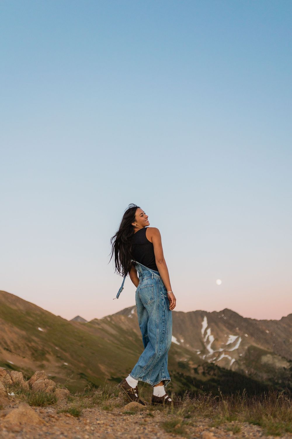 A person in denim overalls dances against a backdrop of mountain peaks and clear blue sky.