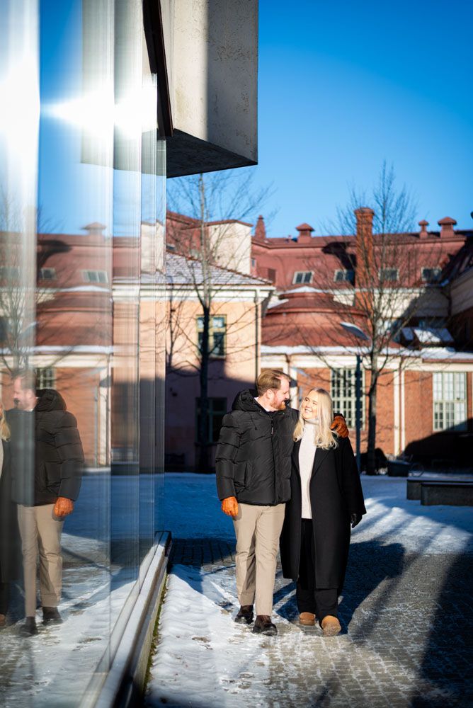 Pariskunta, joka kävelee aurinkoisessa talvisäässä lasiseinän vierellä katsoen toisiaan.