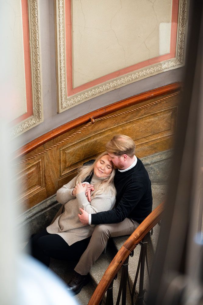 Pariskunta istuu luonnonvaloa täynnä olevassa portaikossa, mies halaa naista ja nainen nojaa mieheen ja katsoo häntä.
