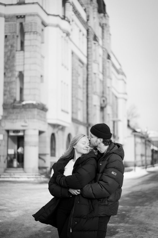 Pariskunta kaupunkimiljöössä. mies halaa vaaleahiuksista naista takaapäin ja suutelee tätä ohimolle.
