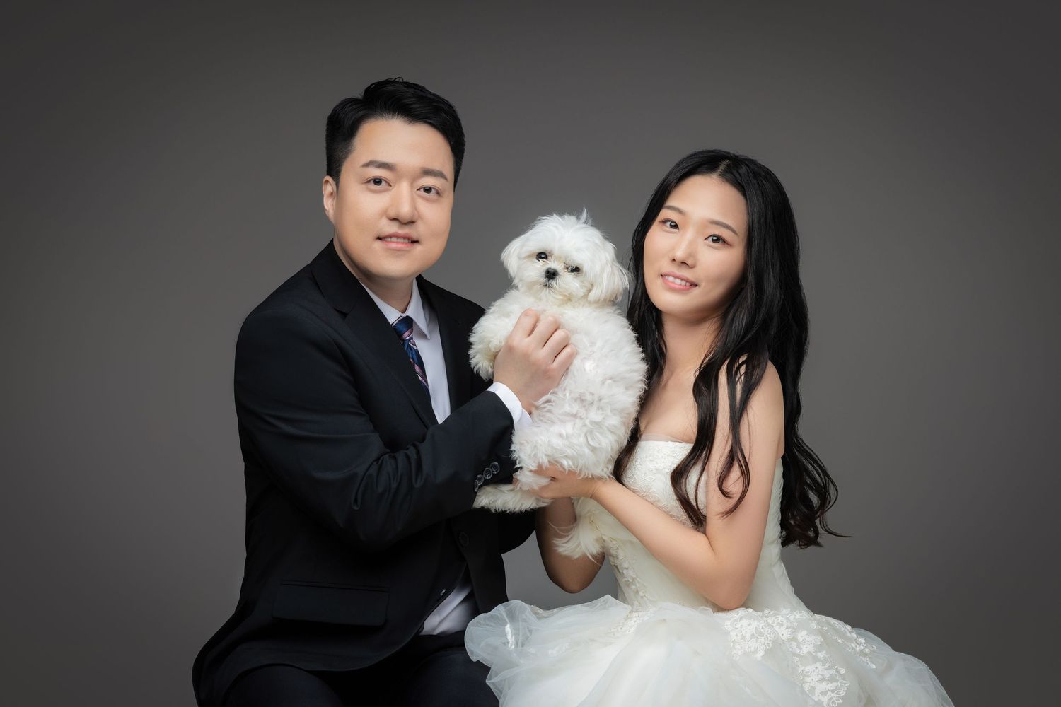 A couple in formal wedding attire poses with a small white dog in a professional studio setting.