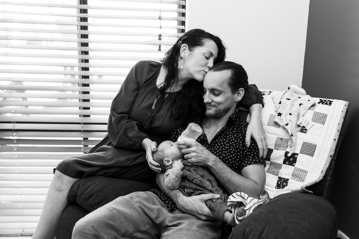 Newborn cuddled with parents at home, photographed naturally with no props or wraps.