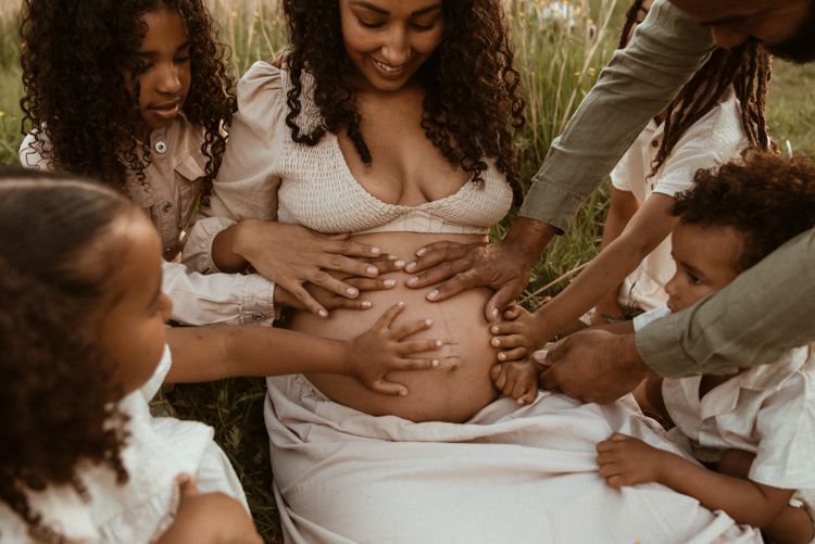 Family members embrace and touch pregnant belly during Manchester maternity photoshoot in rustic outdoor setting.