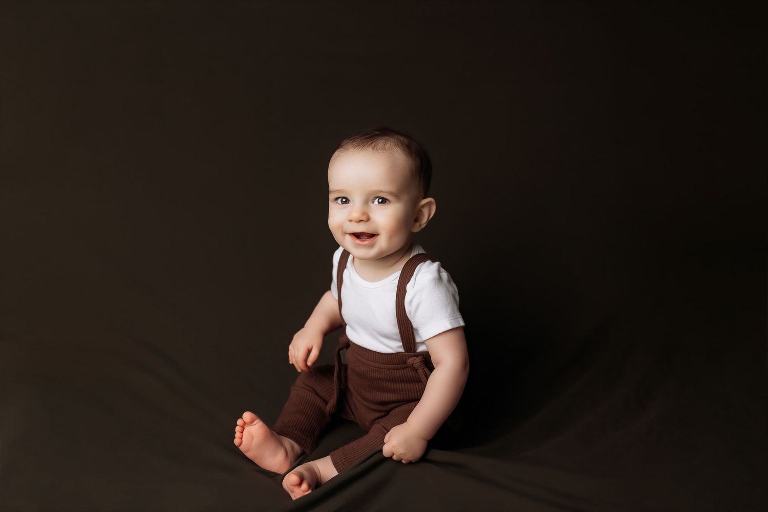 A baby in mint green overalls sits on a wooden floor while playing with cotton balls.
