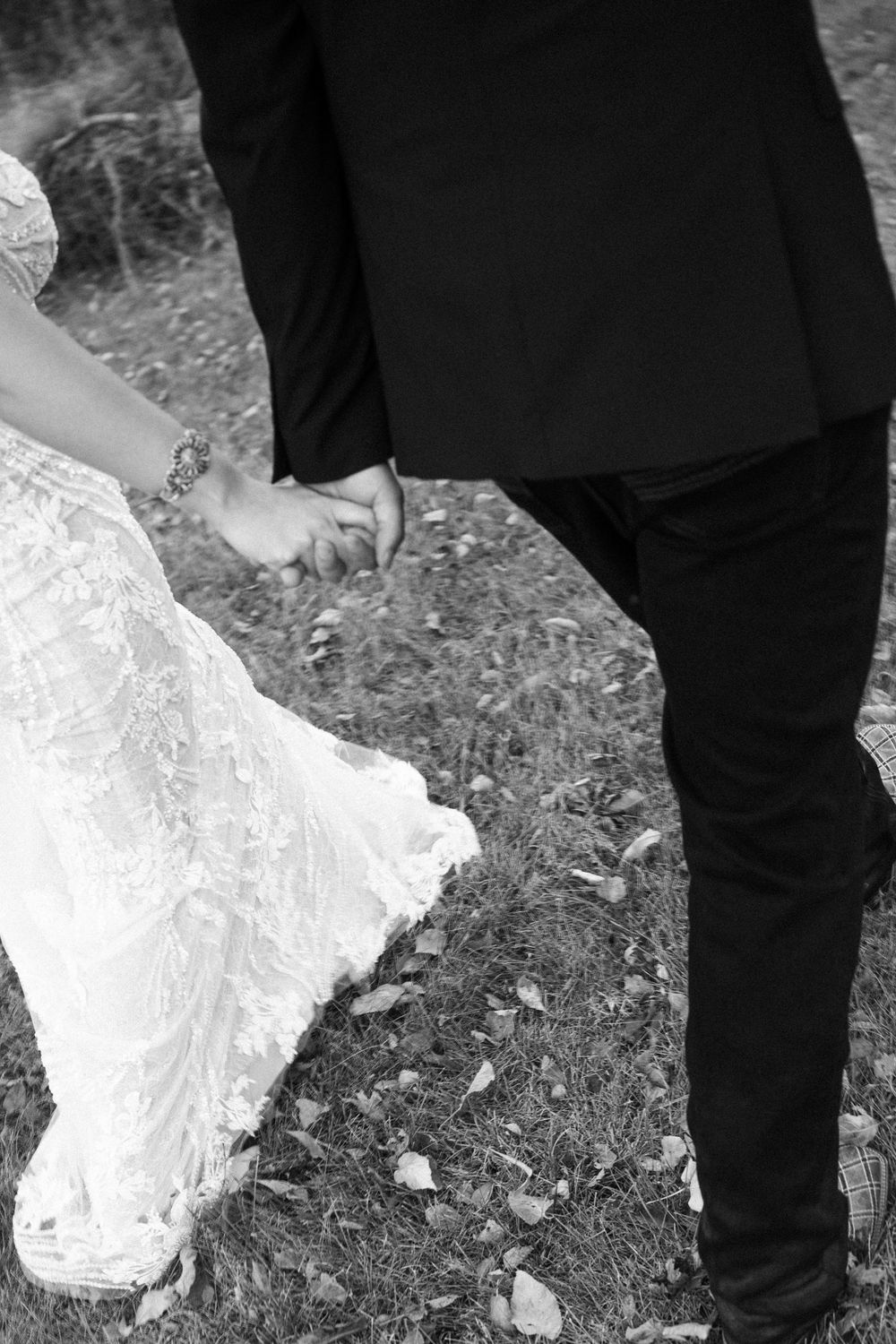 Black and white photo showing a wedding dress hem trailing on rocky ground next to dark pants and shoes.