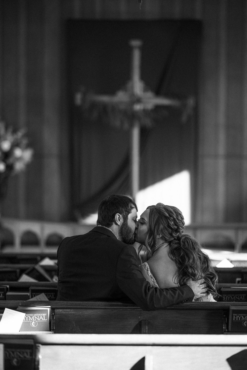 A couple shares a romantic kiss while sitting in a church pew with a cross visible in the background.