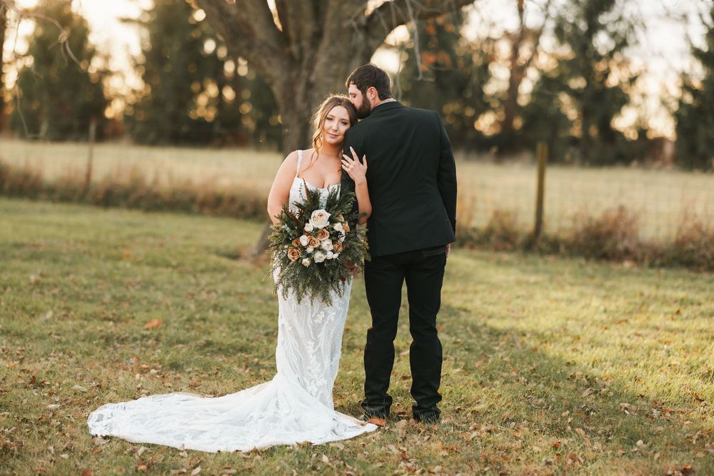 A couple in wedding attire walks together through a golden field during sunset, with the bride's dress trailing behind.