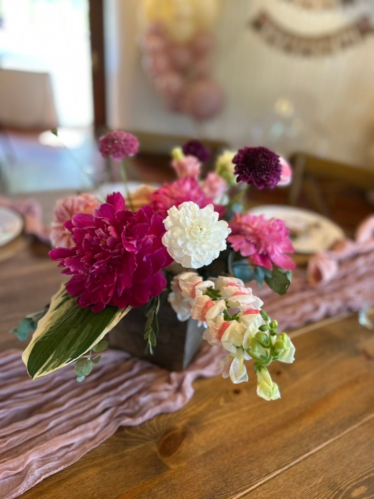 Pink and white floral centerpiece with peonies and dahlias in a dark vase on a dusty rose table runner.