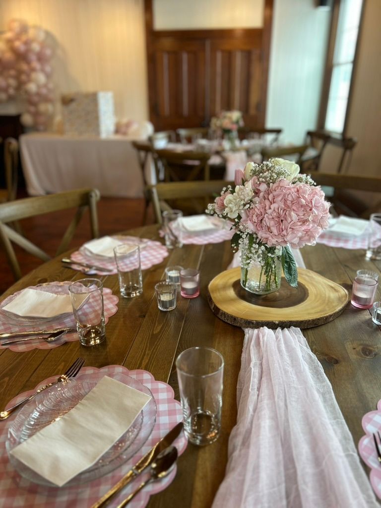 Rustic wooden table with pink gingham placemats, hydrangea centerpiece on wood slice, and candlelit settings.