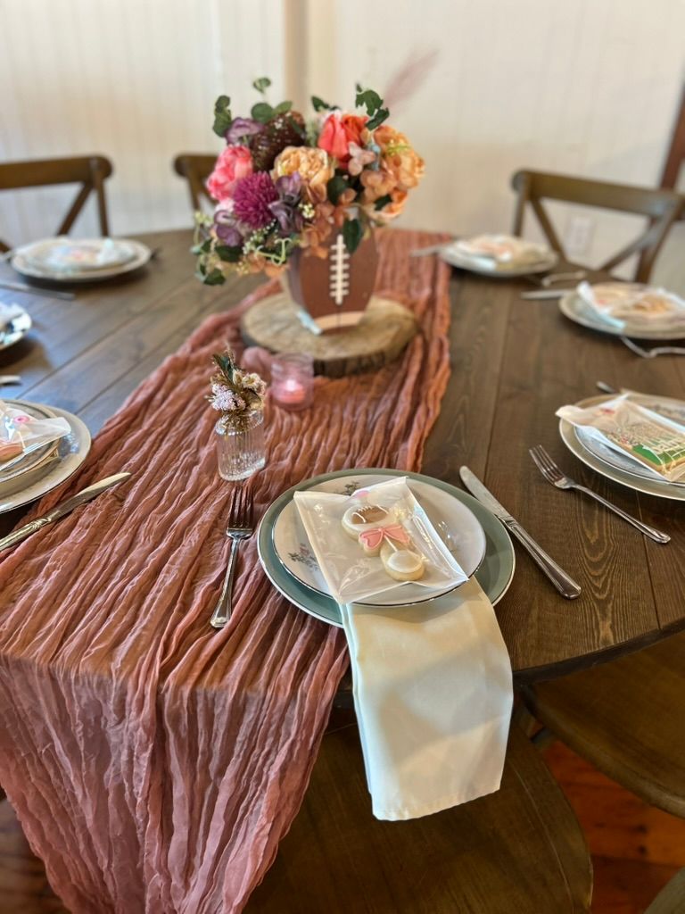 Rustic round table with pink crinkle runner, floral centerpiece in football vase, and elegant place settings.