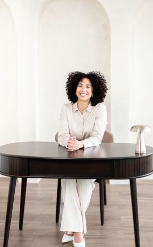 Professional wearing white attire leans on dark wooden table in minimalist studio setting with hardwood floors.