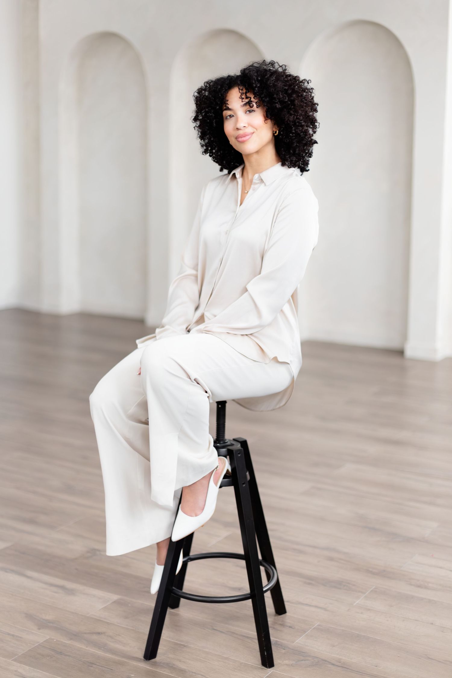 A person in an elegant white outfit poses on a black chair against a minimalist white studio backdrop.