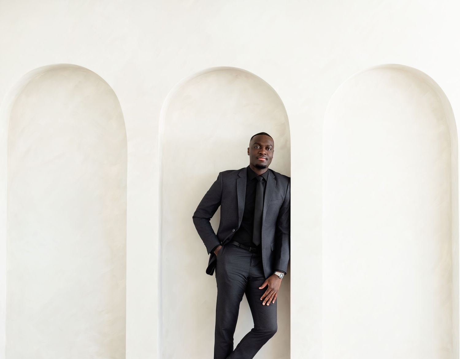A person wearing a sleek black suit poses confidently in a white archway against a minimalist architectural backdrop.