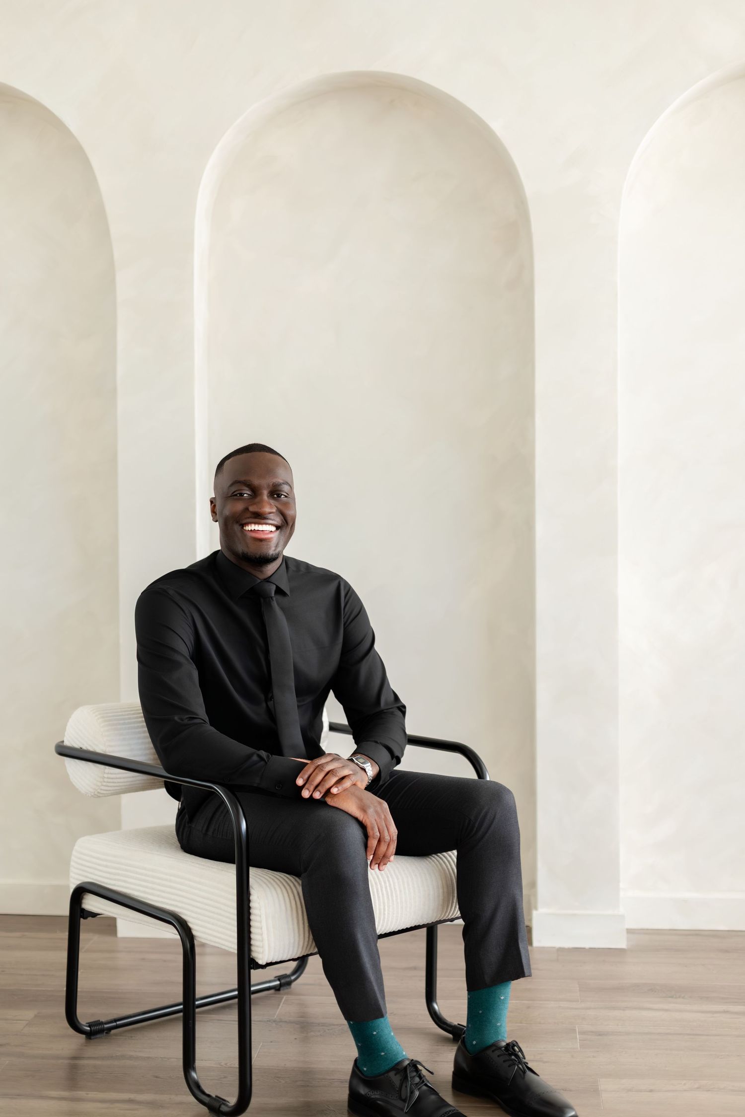 Person in all black attire sits smiling in white chair against arched white wall interior, wearing bright teal socks.