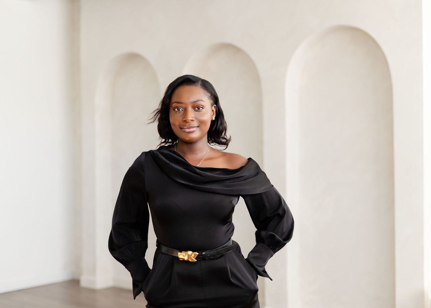 Professional portrait of someone in a black cowl neck dress with long sleeves and gold belt against white arched wall.