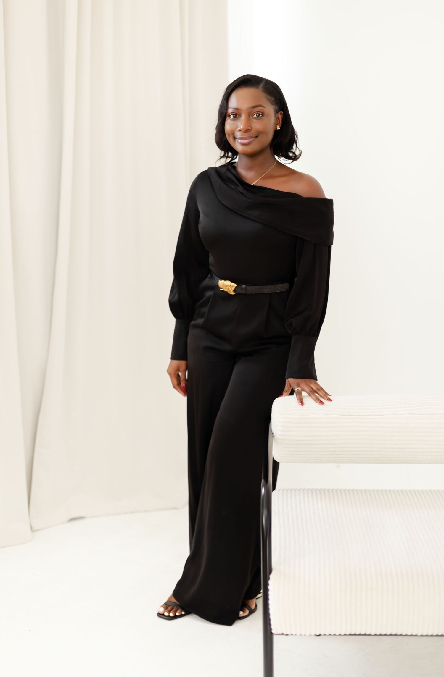 A person wearing an elegant black off-shoulder jumpsuit with long sleeves poses next to a white bench.
