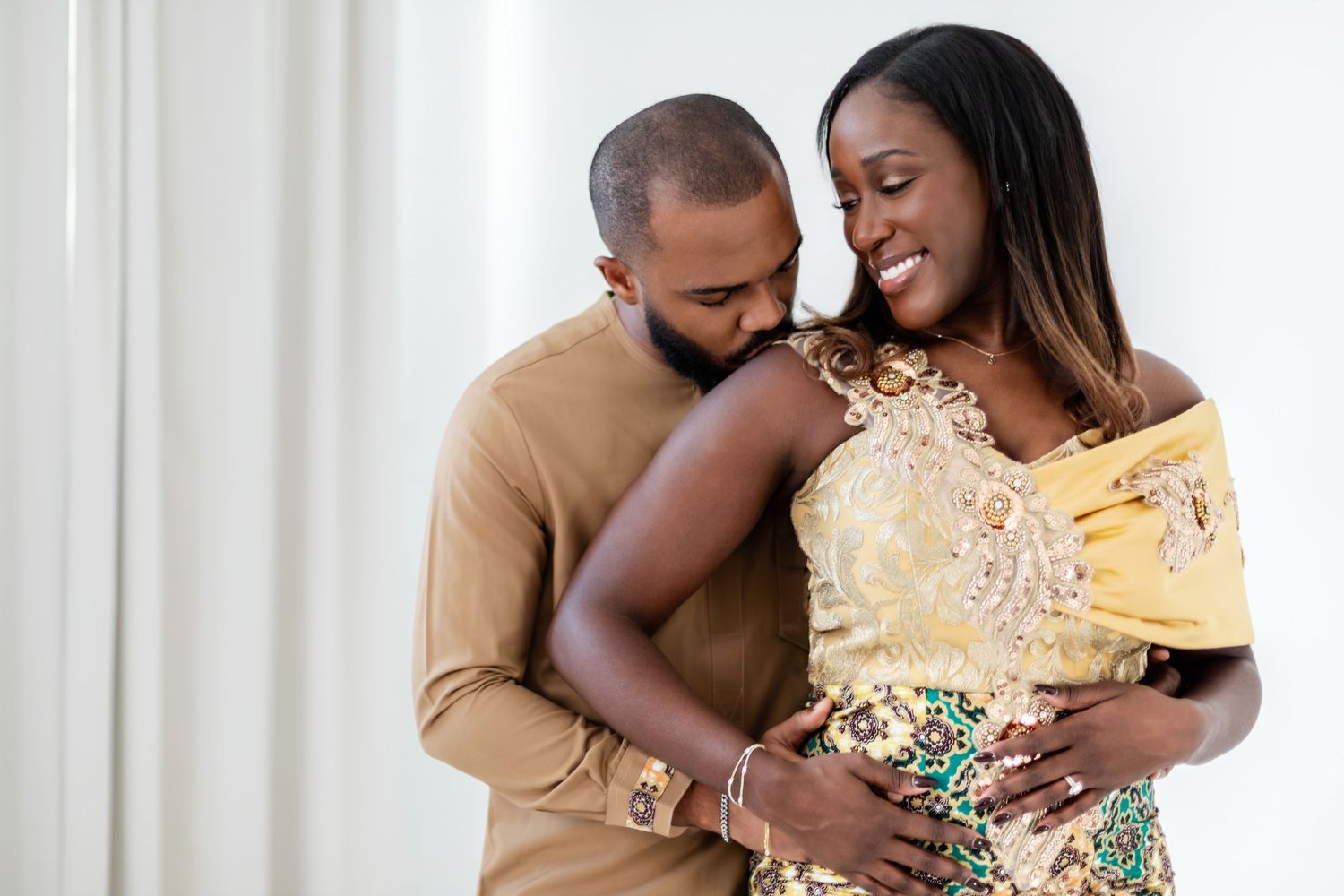 A couple in formal attire share an intimate moment in a white studio setting with arched doorways.