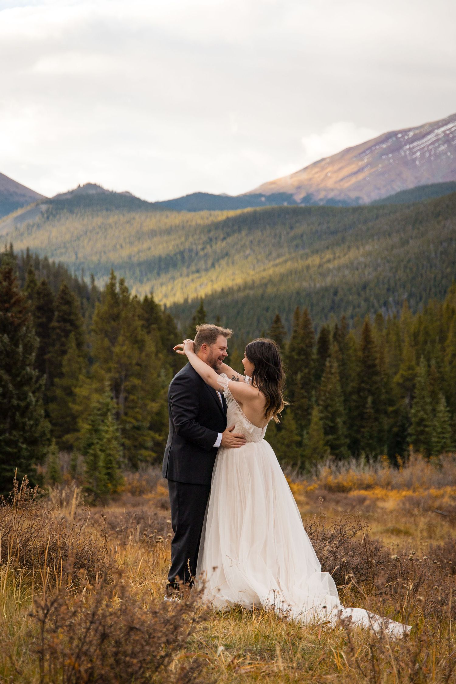 A couple in wedding attire shares a romantic moment against a mountain landscape at sunset.