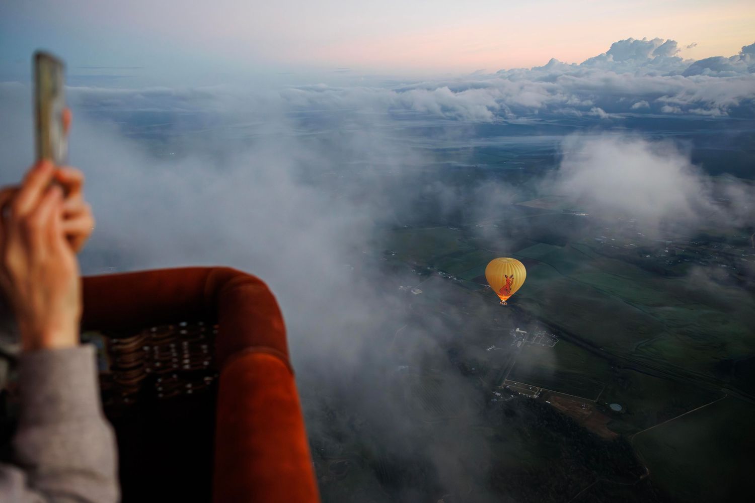 Aerial view from a hot air balloon basket showing another yellow balloon floating above misty green fields at dawn. Cairns Australia