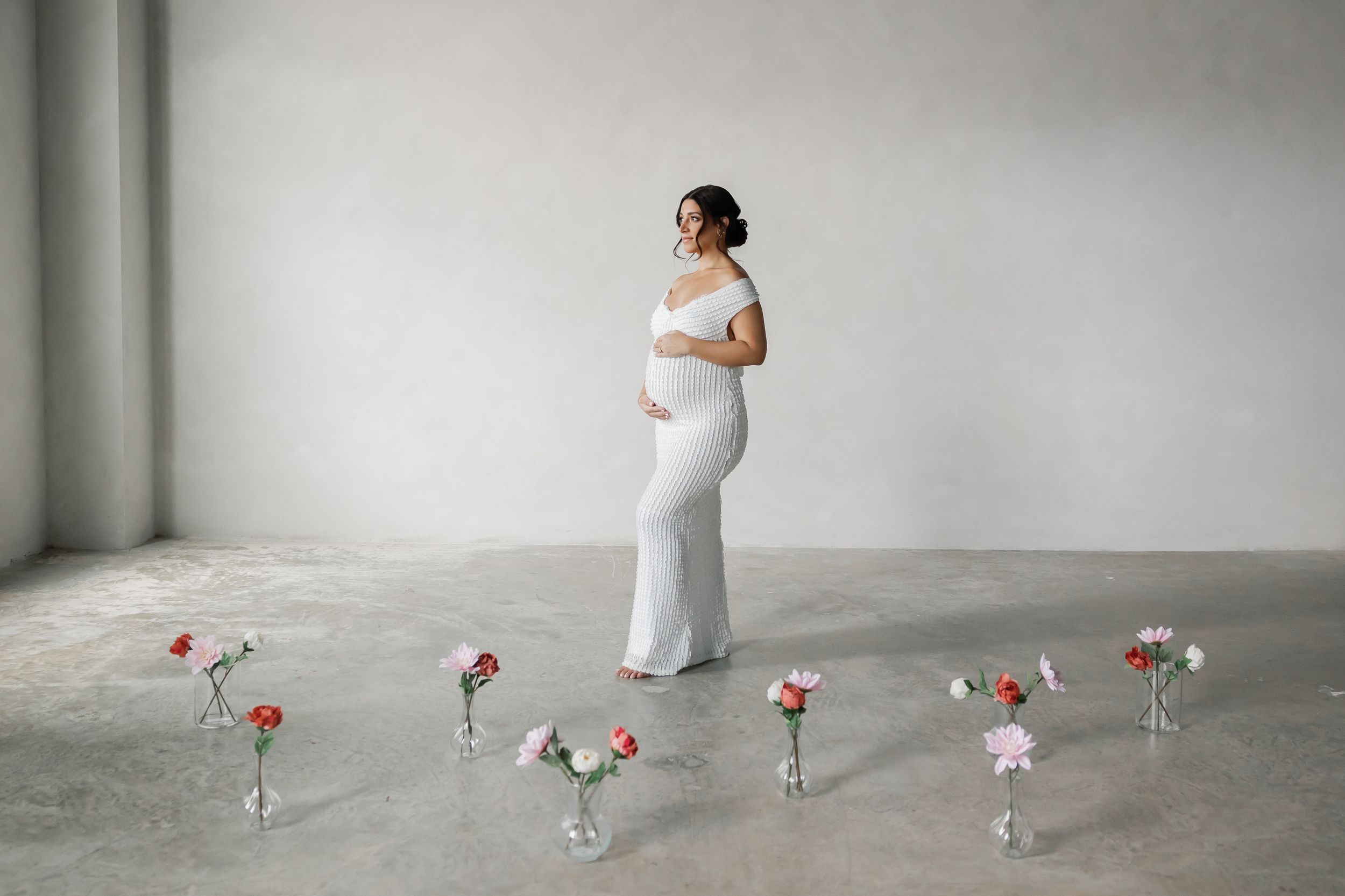 A pregnant woman standing alone in a minimalist, concrete setting with flowers on the floor.