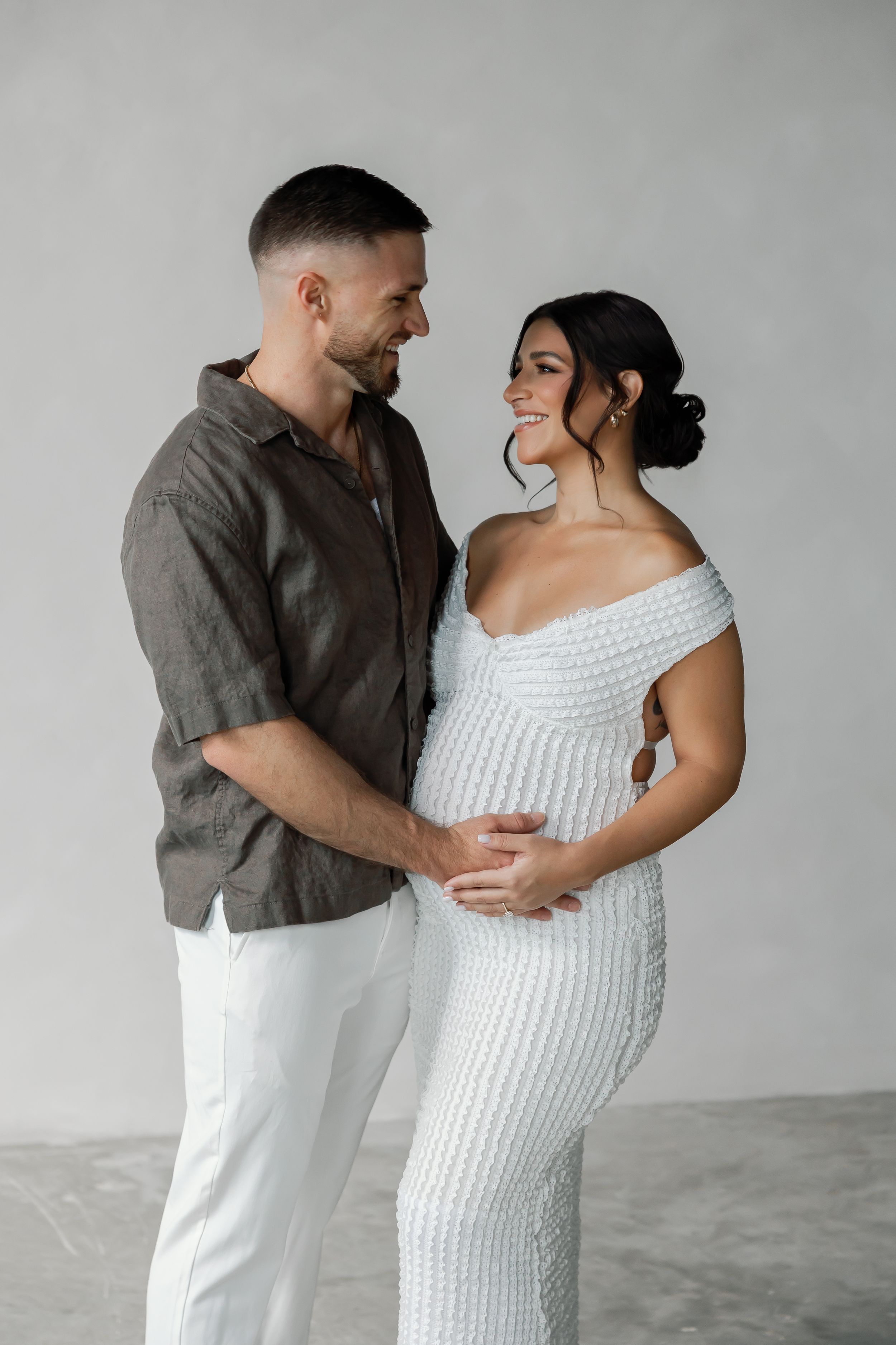 A pregnant woman and a man embracing in a minimalist, concrete setting with flowers on the floor.