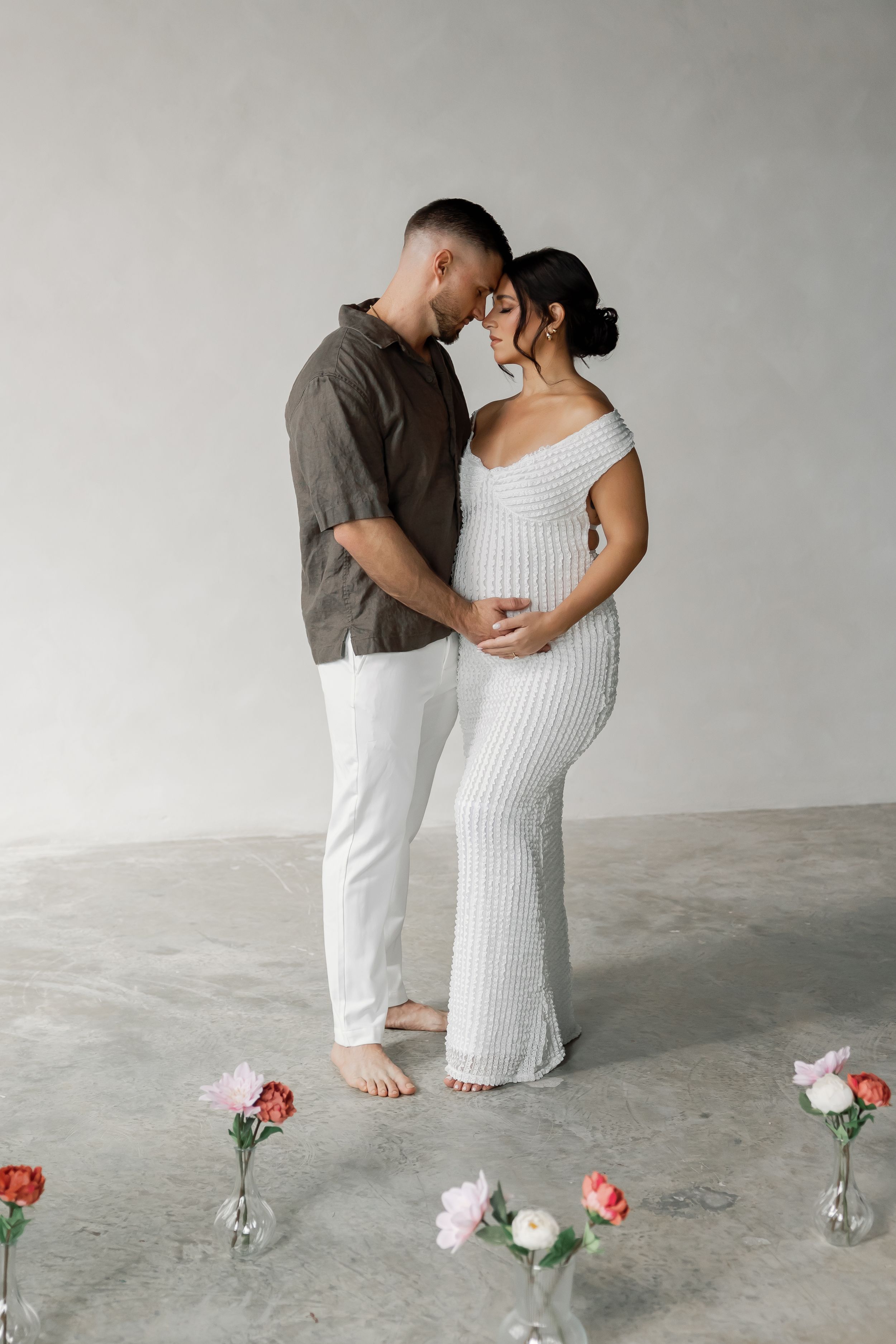A pregnant woman and a man embracing and looking at each other in a minimalist, concrete setting with flowers on the floor.