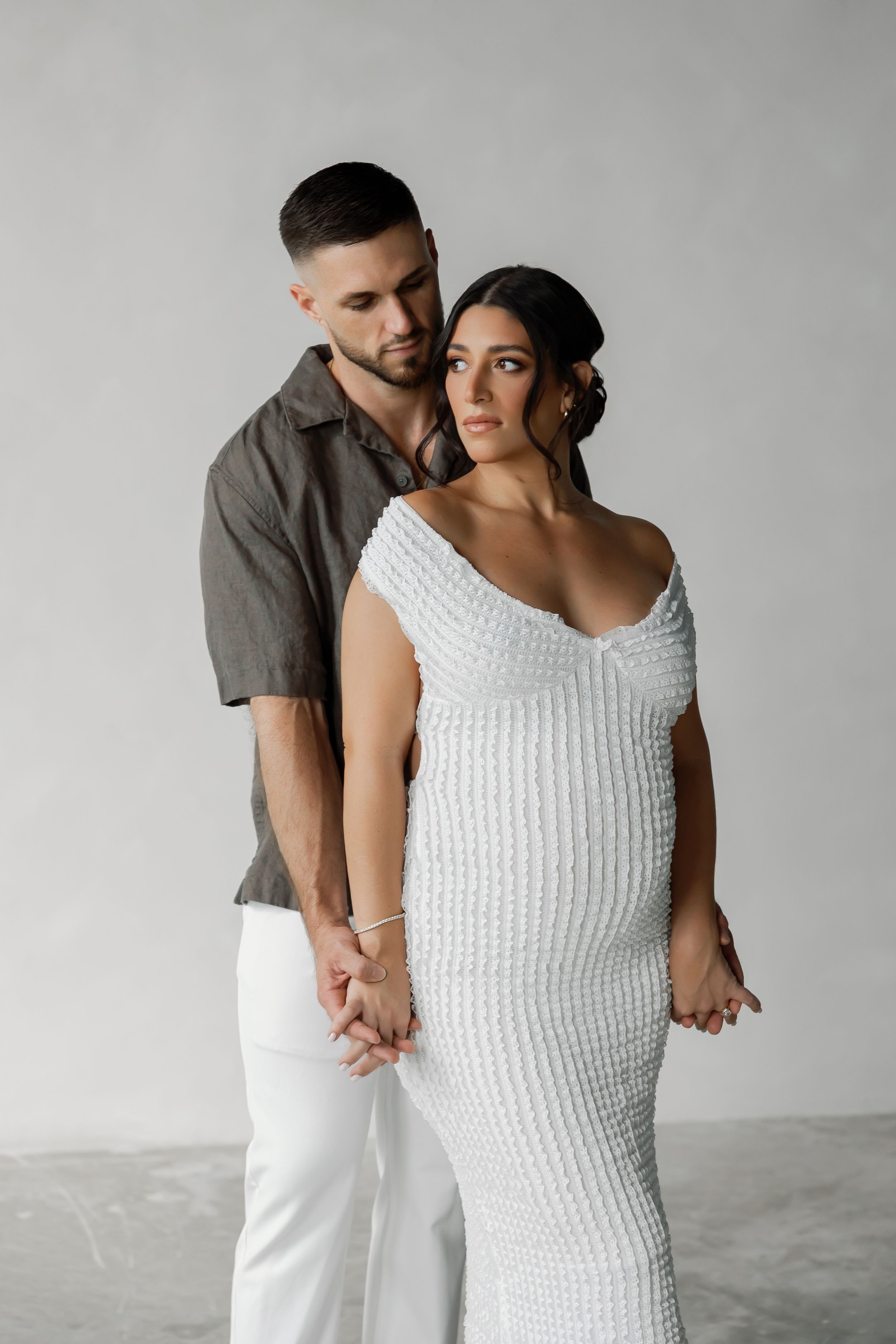 A pregnant woman and a man embracing and looking at each other in a minimalist, concrete setting with flowers on the floor.