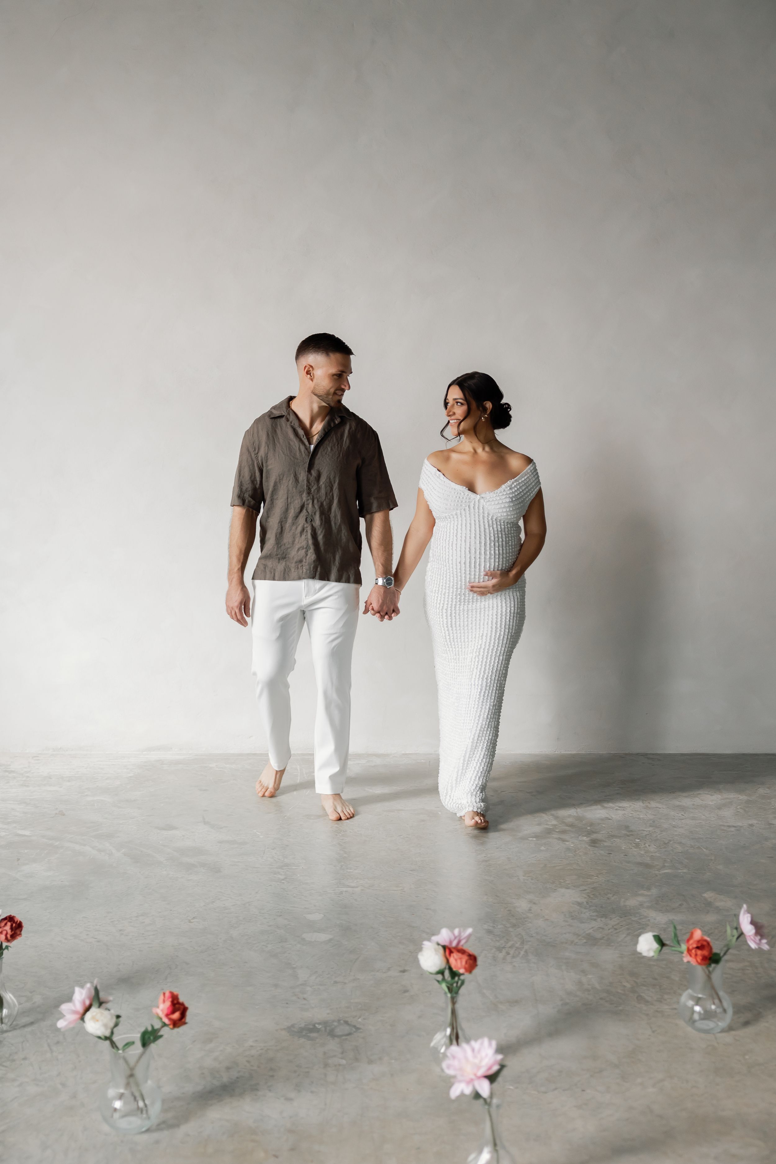 A pregnant woman and a man walking together in a minimalist, concrete setting with flowers on the floor.