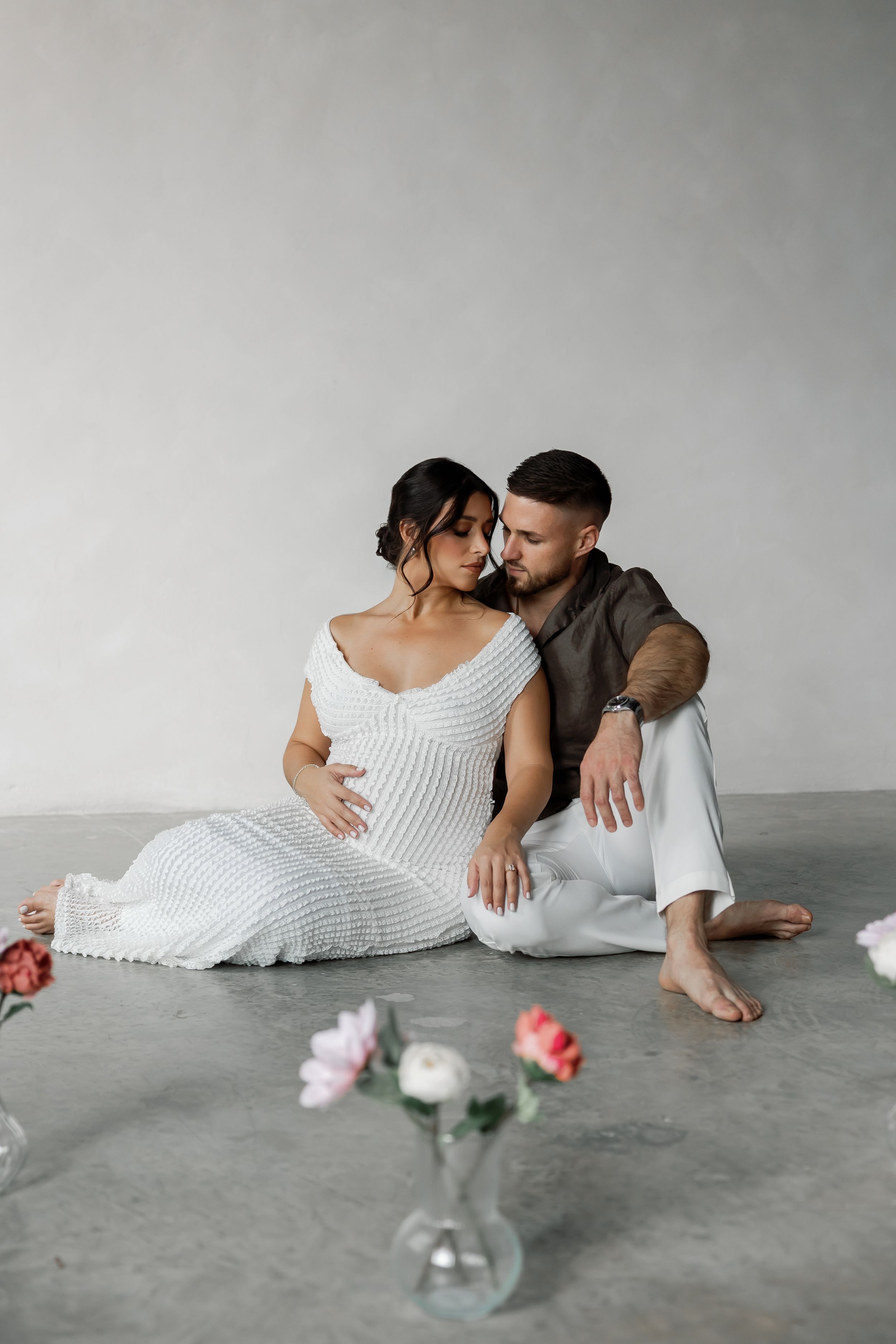 A pregnant woman and a man embracing and looking at each other in a minimalist, concrete setting with flowers on the floor.