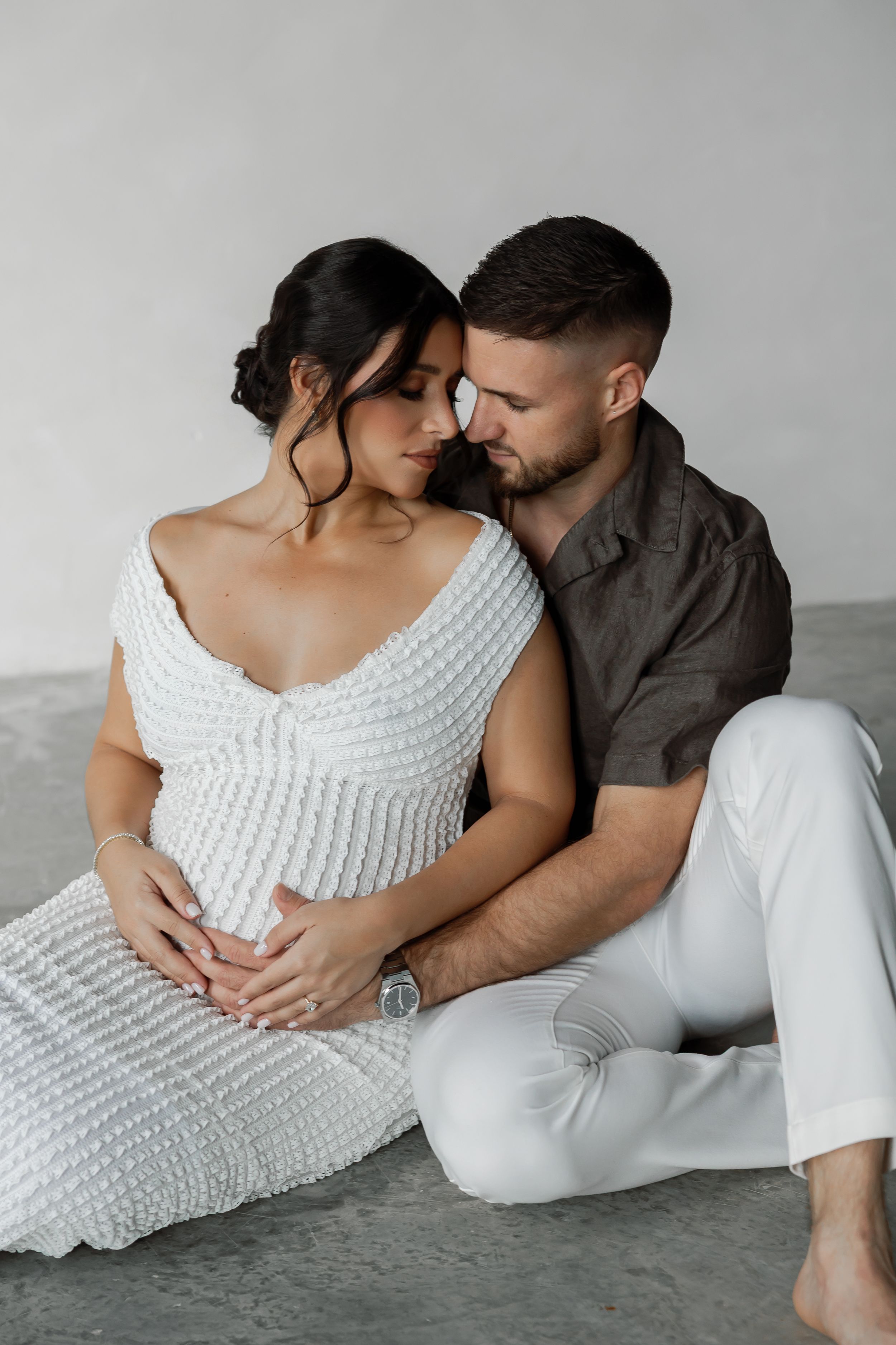 A pregnant woman and a man embracing and looking at each other in a minimalist, concrete setting with flowers on the floor.