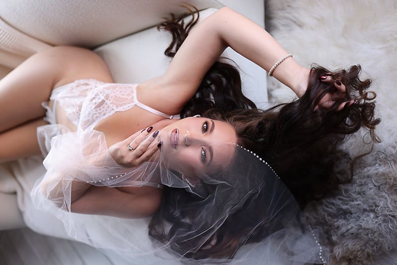 Artistic boudoir photoshoot with white lace lingerie and soft lighting on grey fabric.