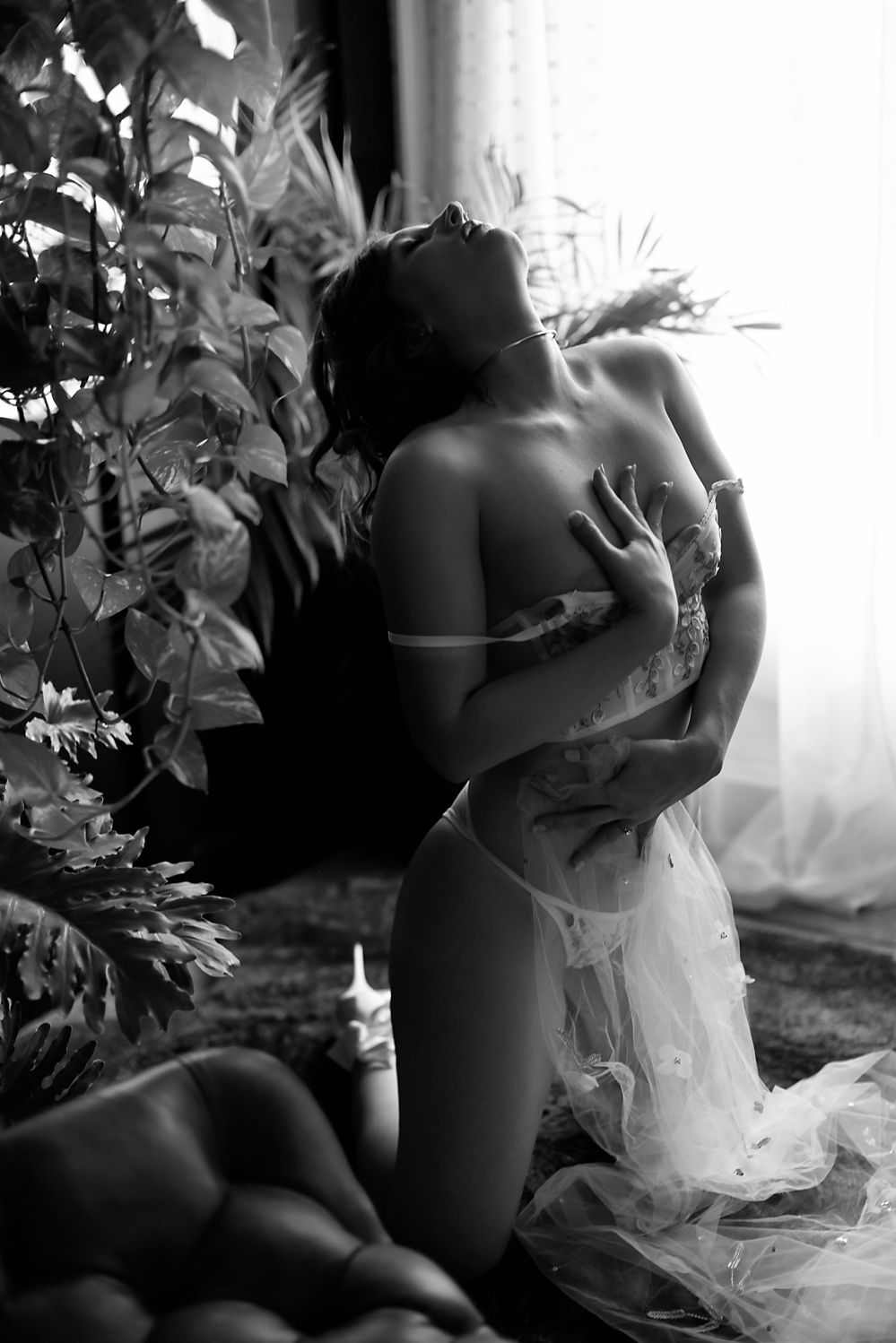 Black and white artistic portrait with sheer fabric and foliage near window.