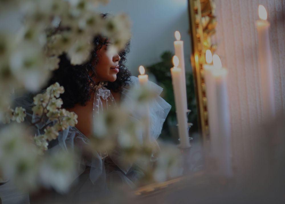 Elegant bride with curly hair sits among white flowers and tall candles in a romantic, softly lit wedding setting.