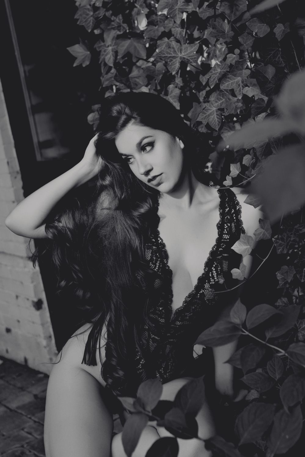 Black and white portrait of woman in lace bodysuit posed against ivy covered wall.