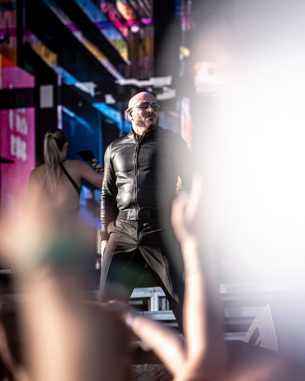 Bald man in black leather outfit performing on a colorful illuminated stage at a live event.