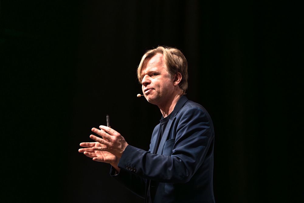 A person in a dark suit gestures while speaking during what appears to be a presentation or speech.