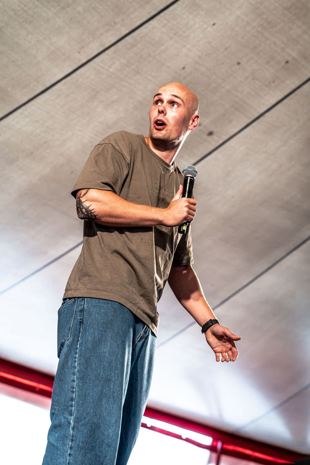 Bald man performing stand-up comedy on stage, holding a microphone in a gray t-shirt and jeans.