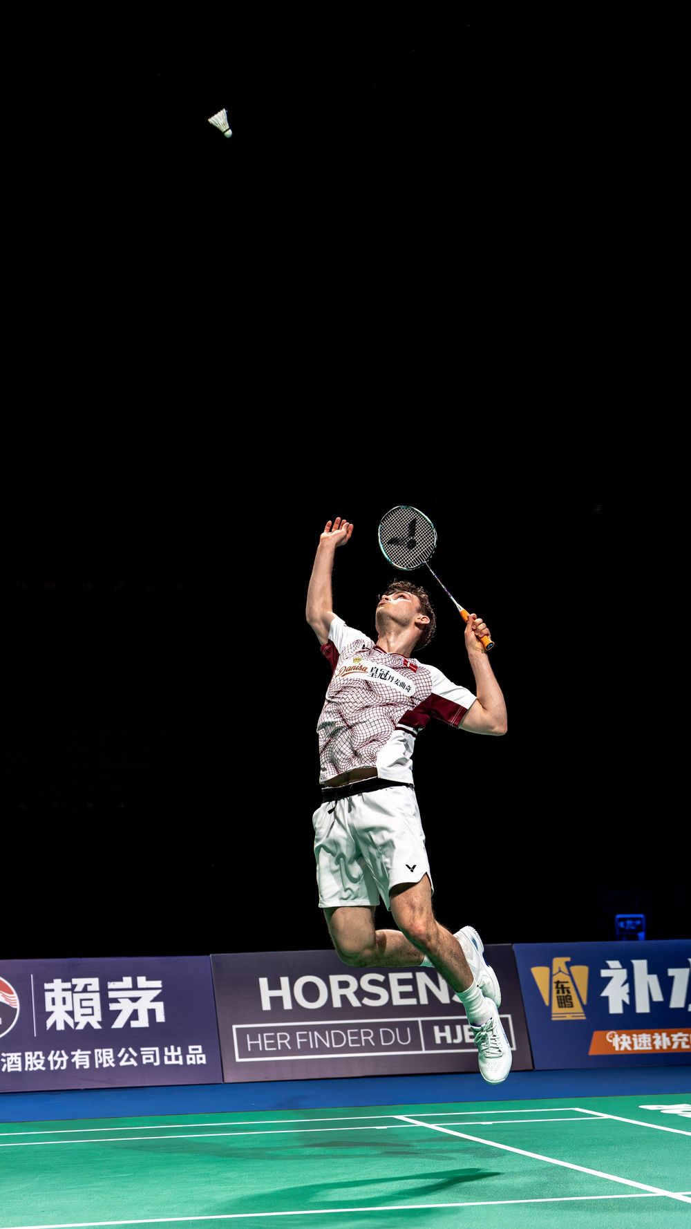 Badminton player leaping mid-air to smash shuttlecock with racket at professional indoor tournament with HORSEN sponsorship banners.