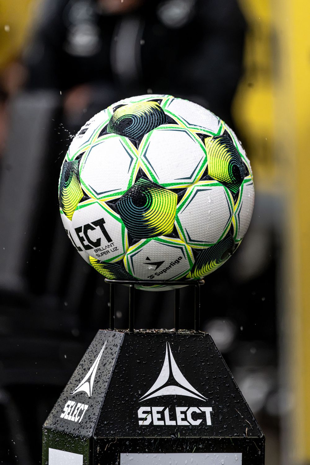 Select branded soccer ball displayed on a pedestal with yellow and green geometric pattern design.