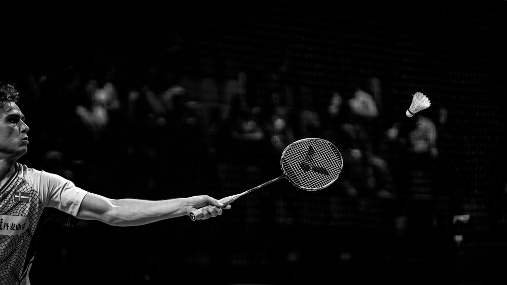 Black and white photo of a badminton player mid-game, reaching to hit a shuttlecock with racket in a packed arena.