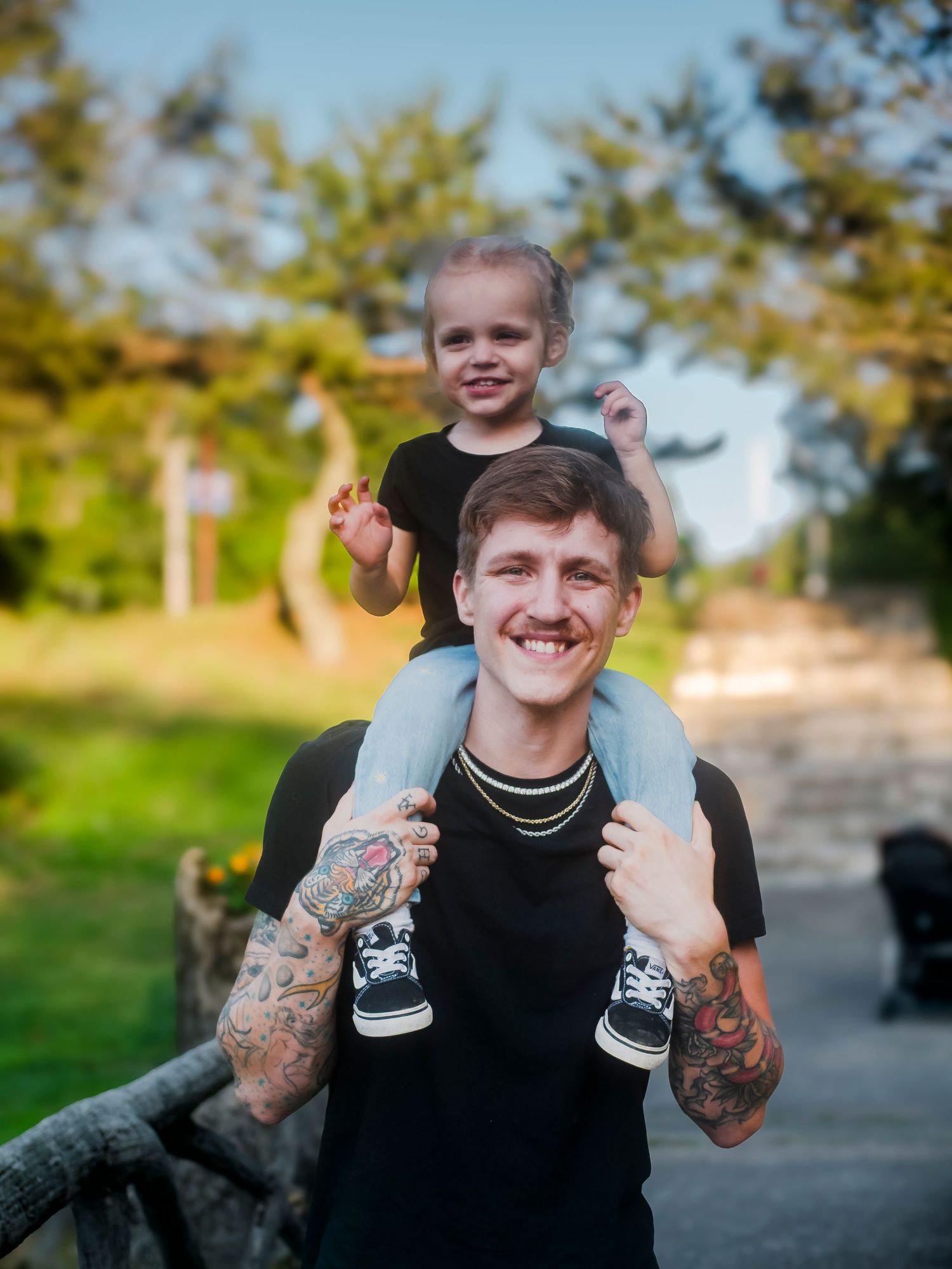A person wearing a black shirt and tattoos carries a child on their shoulders on a sunny day outdoors.