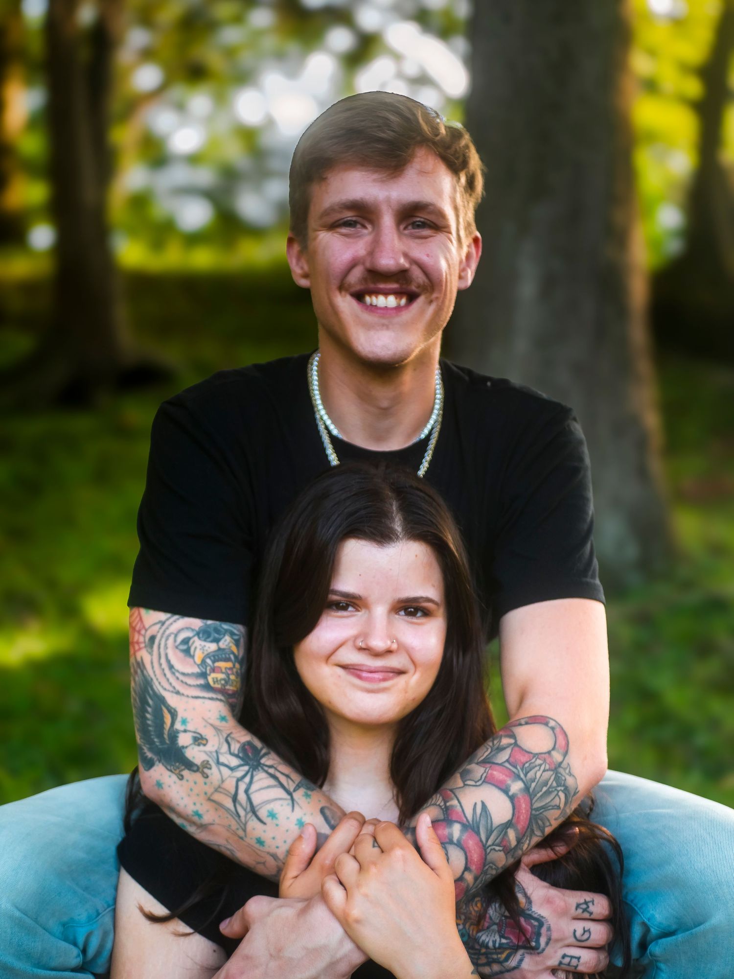 A couple in casual attire share a playful embrace outdoors with visible tattoos and warm smiles in natural lighting.