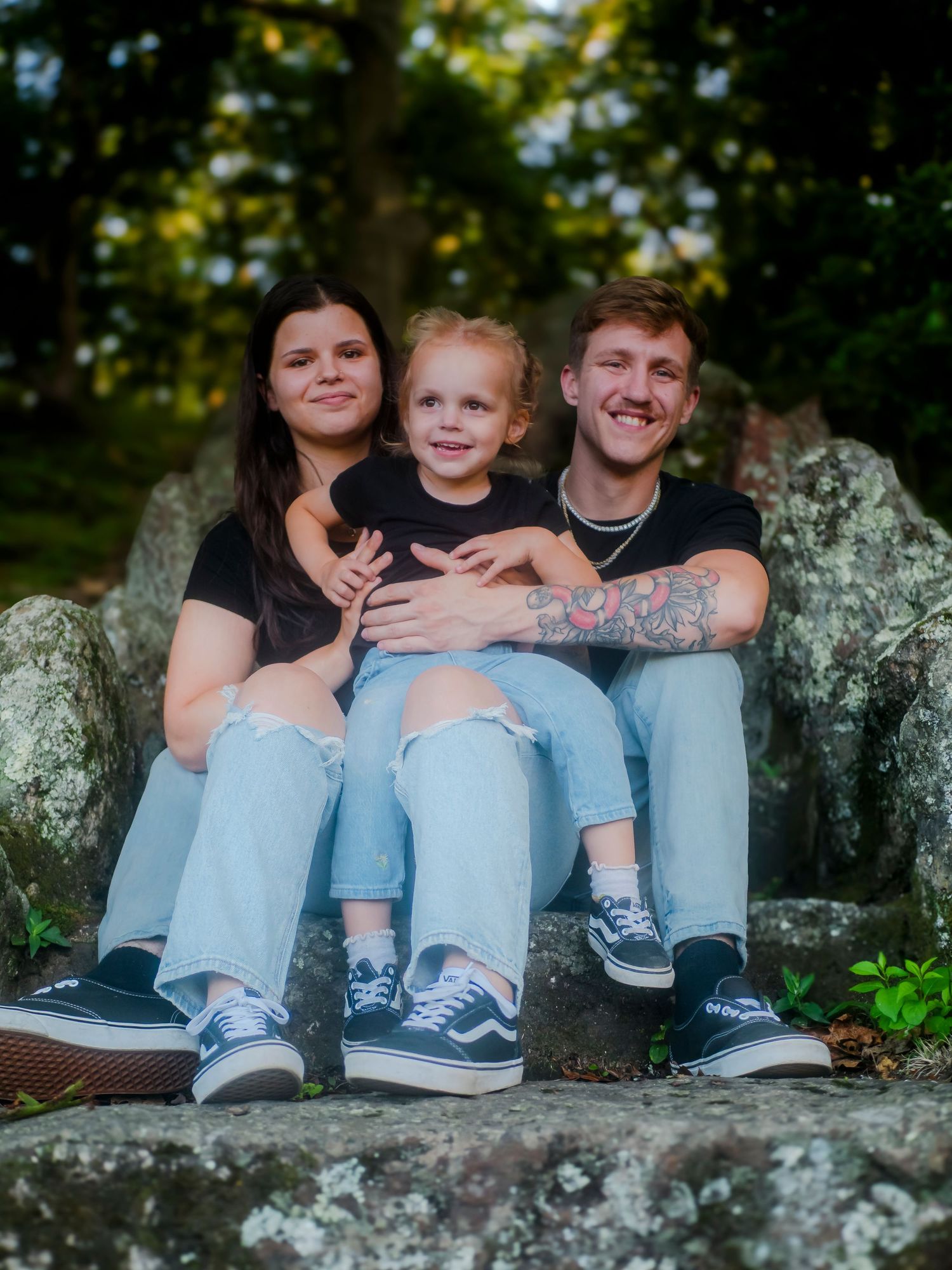 A family sits together on large rocks in a wooded setting wearing casual clothing and sneakers.
